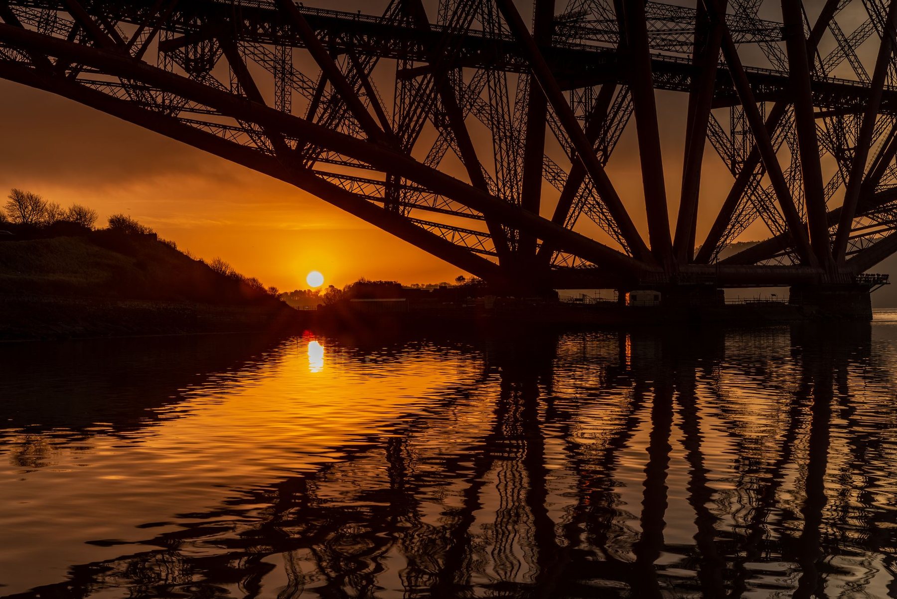The underside of a steel railway bridge at sunset, reflected in still water