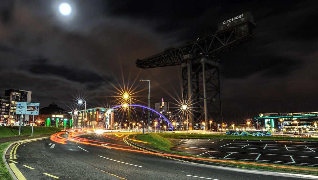 A roadside at night: lit street lamps, road markings, parking bays and a bridge beyond
